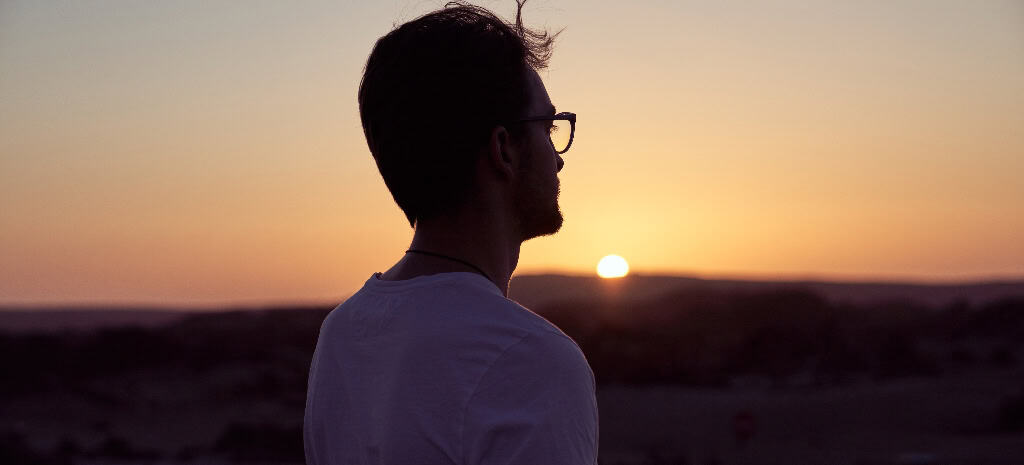 Side view of male traveler in eyeglasses enjoying sunset sky over mountain ridge during trip in nature on summer evening